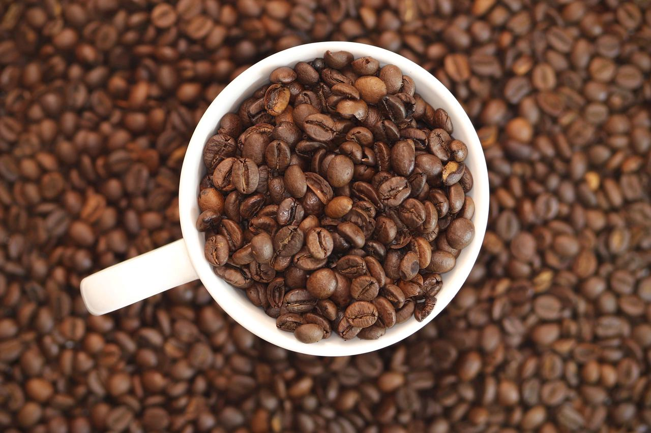 A top view of a white cup filled with dark roasted coffee beans on a pile of coffee beans