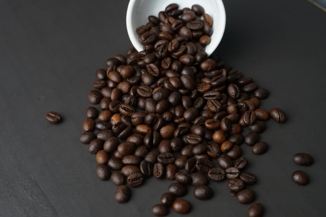 A detailed close-up of roasted coffee beans spilling out of a white cup onto a dark surface