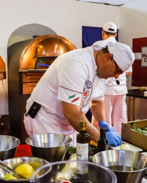 Chef in white uniform preparing ingredients in a professional kitchen with a traditional pizza oven in the background.