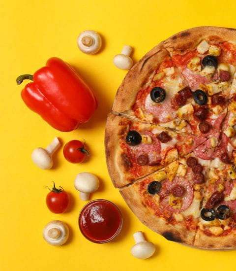 A freshly baked pizza with toppings, surrounded by red pepper, tomatoes, mushrooms, and a small bowl of tomato sauce on a bright yellow background