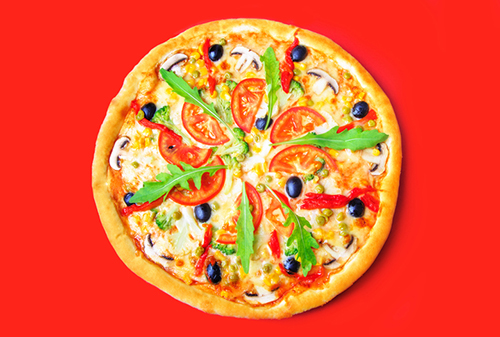 A freshly baked pizza with toppings, surrounded by red pepper, tomatoes, mushrooms, and a small bowl of tomato sauce on a bright yellow background